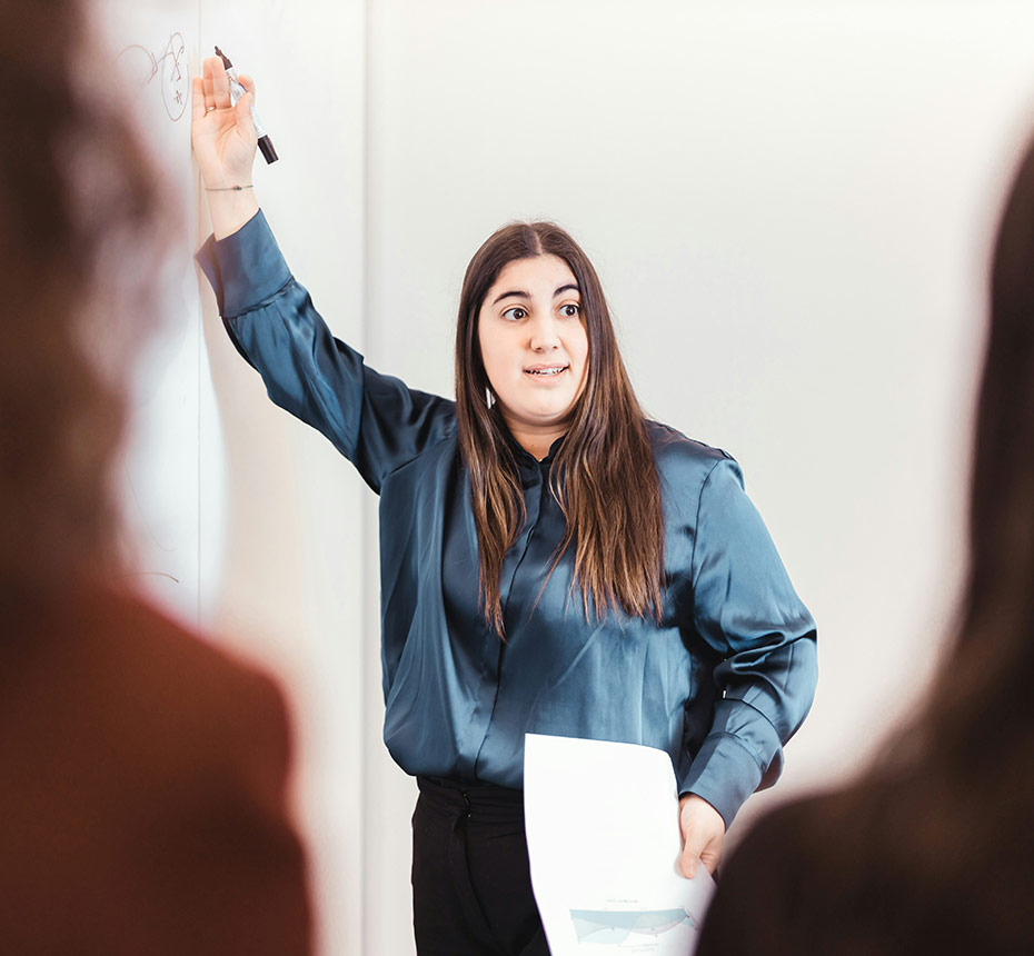 Woman presenting at a whiteboard while holding a paper, speaking to a group.