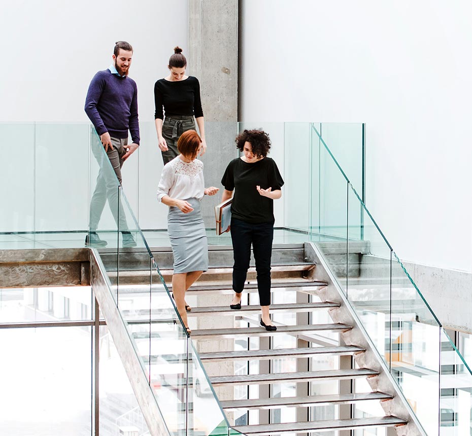Four coworkers walking down glass stairs in a modern office building, engaged in conversation.