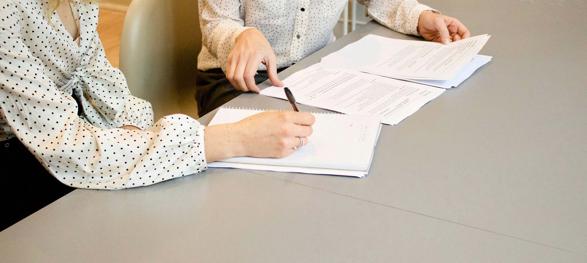 Two women reviewing documents and taking notes at a table during a business or legal meeting.