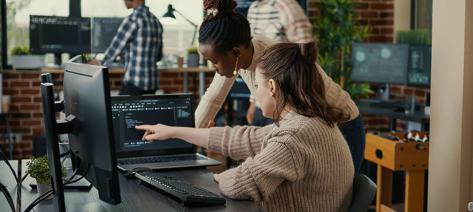 Two women coding together in a tech office with developers in the background.